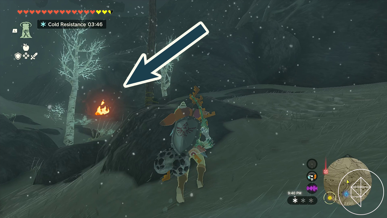 Link walks across a snowy field with ice-covered trees in Zelda: Tears of the Kingdom. A fire burns in the distance near a cave entrance, and an arrow indicates players should move to the fire.